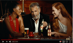 A screenshot from a Dos Equis commercial of the same stylish man. He is sitting in between two attractive young women in a bar holding a Dos Equis Beer in his hand and posing into the camera with a confident look.