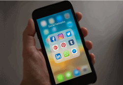 A hand holding an iPhone showing various social media apps