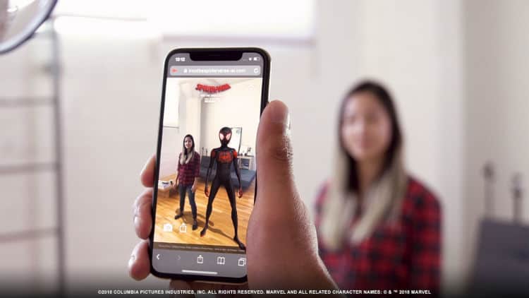 Person holding smartphone which uses augmented reality technology to project Spider-Man next to a woman standing in the background.