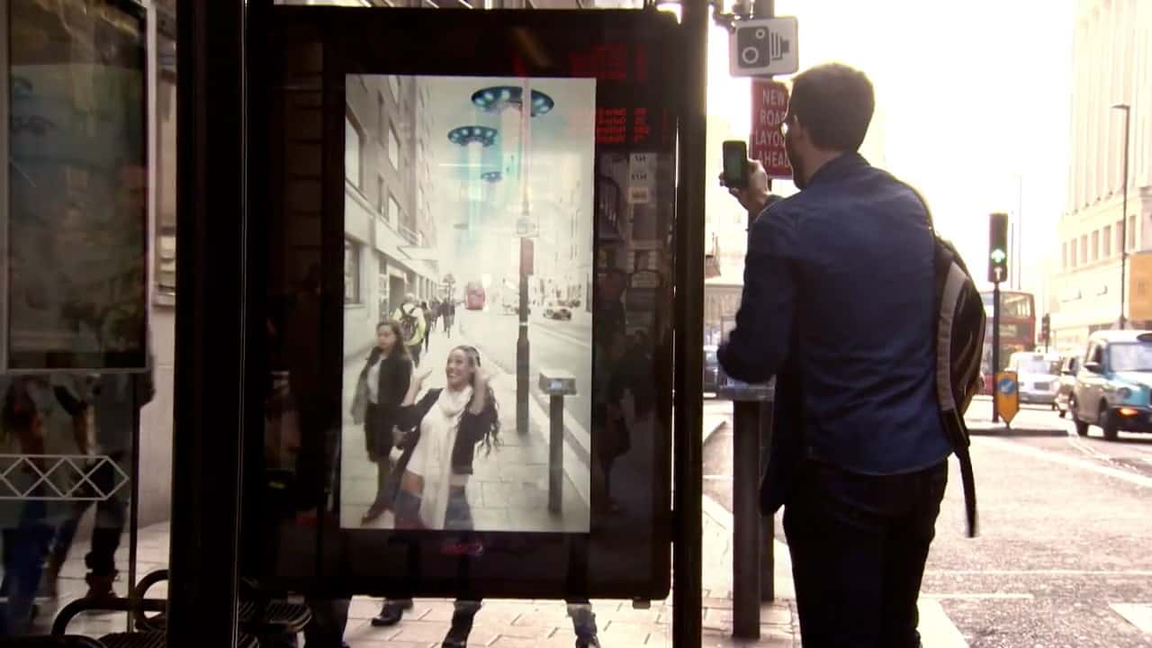 Man takes a photo with his smartphone of the augmented reality screen installed in the bus shelter. On the screen, two UFOs fly above passers-by.
